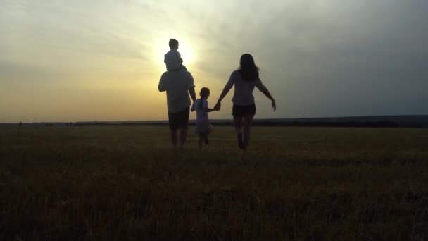 Feliz silueta de paseo familiar al atardecer. Mamá, papá, hijo e hija caminan tomados de la mano en el campo. — Vídeos de Stock