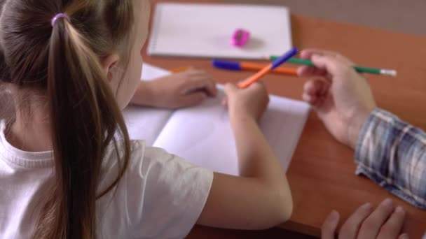 Een klein meisje met een leraar zit aan een schoolbureau en tekent in een notitieboekje met een pen.4k — Stockvideo