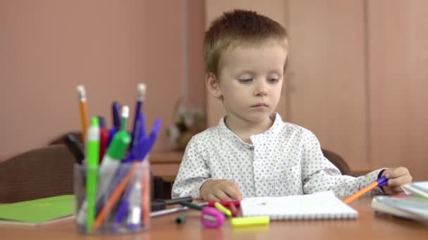 Um menino bonito de três anos senta-se em sua mesa na classe e pensa sobre a resposta. — Vídeo de Stock