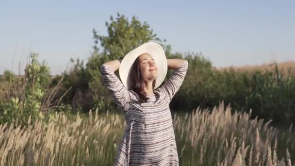 Belle jeune fille dans un chapeau de paille. Elle se tient dans un champ et regarde le coucher du soleil. — Video