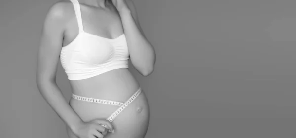 Vacker gravid ung kvinna i kläder för gravida kvinnor mäter hennes nakna mage, leende, på en biege bakgrund. Bild på lycklig gravid kvinna som poserar över muren. Titta på kameran — Stockfoto