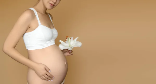 Mooie zwangere jonge vrouw in kleding voor zwangere vrouwen is het meten van haar blote buik, glimlachen, op een biege achtergrond. Foto van een gelukkige zwangere vrouw die over de muur poseert. Kijkend naar camera — Stockfoto