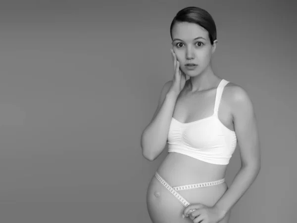 Красива вагітна молода жінка в одязі для вагітних вимірює її голий животик, посміхаючись, на фоні. Зображення щасливої вагітної жінки, що позує над стіною. Дивлячись на камеру — стокове фото