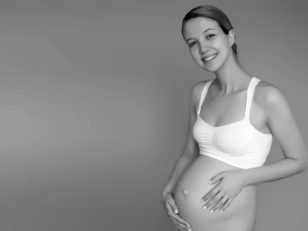 Schöne schwangere junge Frau in Kleidung für Schwangere misst ihren nackten Bauch, lächelnd, auf einem Hintergrund. Bild einer glücklichen Schwangeren, die über einer Mauer posiert. Blick in die Kamera — Stockfoto