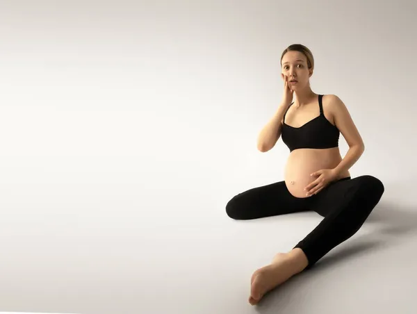Gravid leende kvinna sitter på vitt golv i studiorummet. Titta på kameran. Moderskap. Moderskap. Ung lycklig gravid kvinna sitter. Överraskad Gravid kvinna. Människobegreppet — Stockfoto