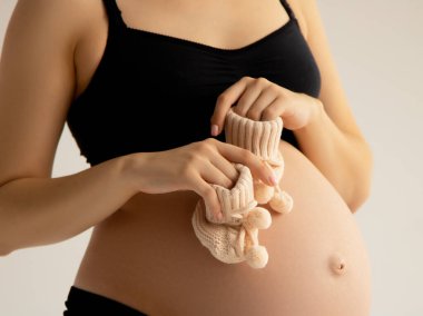 Hamile bir kadının karnındaki doğmamış bebek için küçük ayakkabılar. Kapatın. Annelik, hamilelik, insanlar ve beklenti konsepti. Hamile bir kadın bebek bekliyor. Beyaz arka planı olan güzel bir kadın.