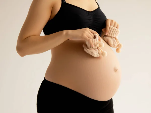 Kleine Schuhe für das Ungeborene im Bauch einer Schwangeren. Aus nächster Nähe. Mutterschaft, Schwangerschaft, Menschen und Erwartungskonzept. Schwangere erwartet Baby. Schönheit Frau mit auf weißem Hintergrund — Stockfoto