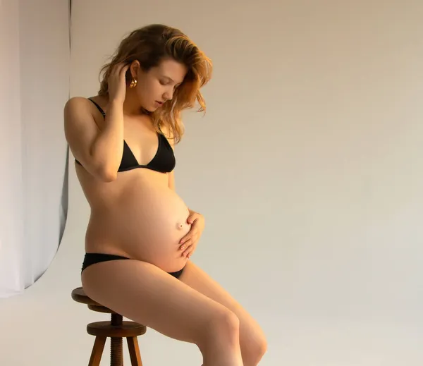 妊娠中のお母さんでお腹が膨れてる。スタイリッシュなマザーフードのコンセプト。スタイリッシュな黒のブラジャーと現代のイヤリングで幸せな若い妊婦は、腹のバンプを保持し、光の中でポーズ。ファッション写真 — ストック写真