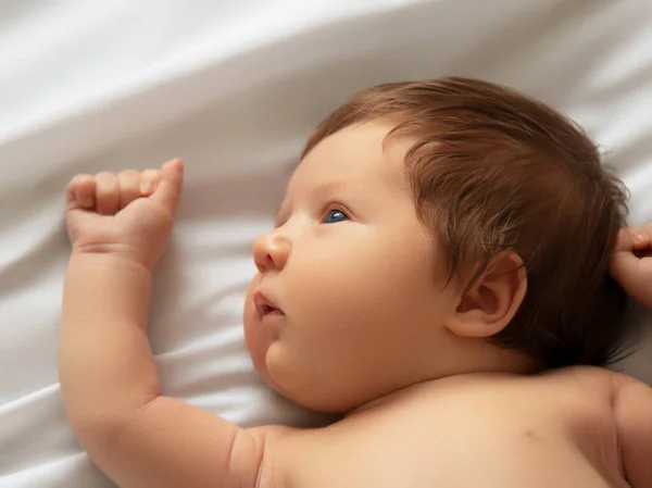 Ładny nowonarodzony chłopiec, portret z bliska. Leży w białym łóżku. Cute noworodka 1 miesiąc życia na białym prześcieradle w pieluszce. Pielęgnacja noworodków, kolka, zęby, noworodek — Zdjęcie stockowe