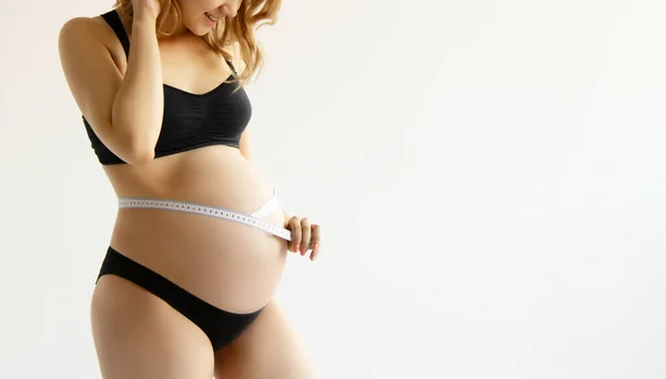 Vacker gravid ung kvinna i kläder för gravida kvinnor mäter hennes nakna mage, leende, på en biege bakgrund. Leende gravid kvinna mäter hennes stora mage — Stockfoto