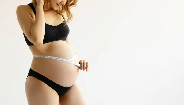 Belle jeune femme enceinte en vêtements pour femmes enceintes mesure son ventre nu, souriant, sur un fond de siège. Femme enceinte souriante mesurant son gros ventre Images De Stock Libres De Droits