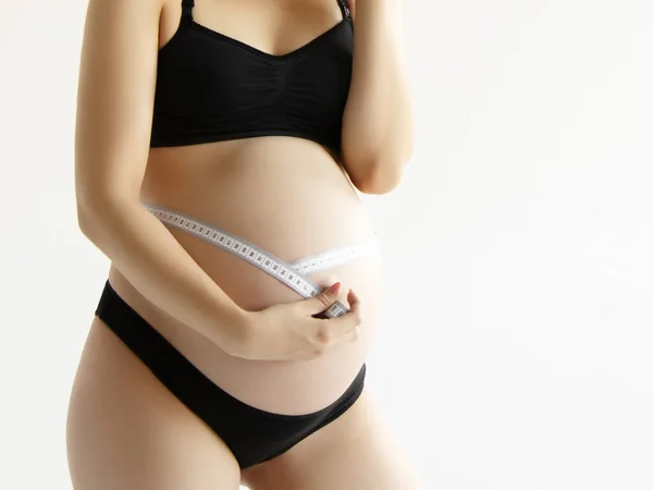 Красива вагітна молода жінка в одязі для вагітних вимірює її голий животик, посміхаючись, на білому тлі. Усміхнена вагітна жінка вимірює великий живіт — стокове фото
