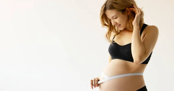 Schöne schwangere junge Frau in Kleidung für Schwangere misst ihren nackten Bauch, lächelnd, auf einem biegen Hintergrund. Lächelnde Schwangere misst ihren großen Bauch lizenzfreie Stockbilder