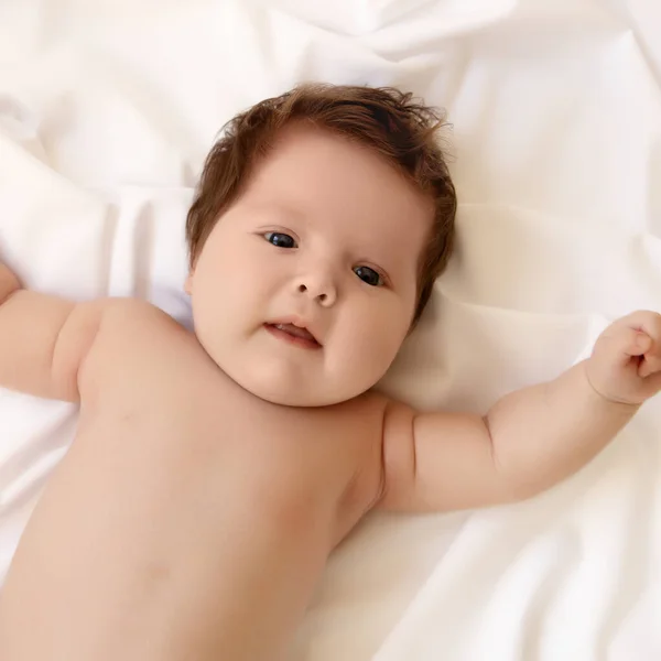 Симпатичная годовалая девочка на кровати на животе с поднятой головой смотрит в камеру большими глазами. Естественное освещение спальни. Младенец. Здоровая и медицинская концепция. Счастливой беременности. — стоковое фото