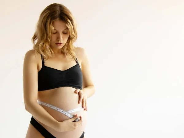 Vacker gravid ung kvinna i kläder för gravida kvinnor mäter hennes nakna mage, leende, på en vit bakgrund. Leende gravid kvinna mäter hennes stora mage — Stockfoto