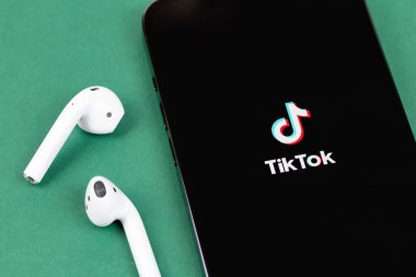 TikTok logo mobile app on screen smartphone iPhone with AirPods headphones on green background. TikTok is app to create and share videos. Moscow, Russia -  July 27, 2021