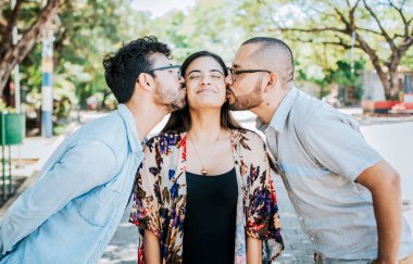 Love triangle concept. Polygamy concept. Two men kissing a girl cheek. Portrait of two guys kissing a girl cheek. Two young men kissing a woman cheek outdoor clipart