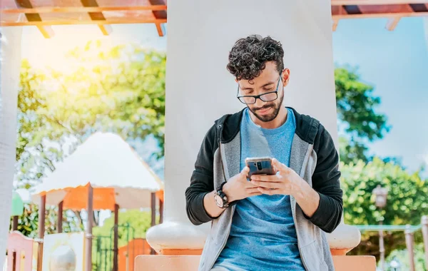 Handsome guy in glasses leaning on a wall chatting on his cell phone, Teenage male leaning on a wall sending a text message, Smiling handsome man using a phone leaning on a wall in a park