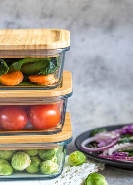 Cam kaplarda çeşitli sebzeler: havuç, ıspanak, domates, Brüksel lahanası. Konteynırlarda vejetaryen yemeği ve atıştırmalıklar, gri arka plan. Temiz yemek, çiğ yemek, detoks, bitki temelli diyet konsepti.