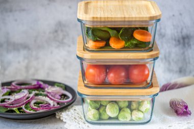 Cam kaplarda çeşitli sebzeler: havuç, ıspanak, domates, Brüksel lahanası. Konteynırlarda vejetaryen yemeği ve atıştırmalıklar, gri arka plan. Temiz yemek, çiğ yemek, detoks, bitki temelli diyet konsepti.