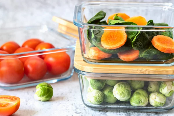 Various vegetables in glass containers: carrots, spinach, tomatoes, Brussels sprouts. Vegan food and snacks in containers, gray background. Clean eating, raw food, detox, plant based diet concept.