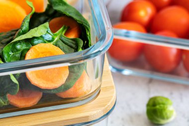 Cam kaplarda çeşitli sebzeler: havuç, ıspanak, domates, Brüksel lahanası. Konteynırlarda vejetaryen yemeği ve atıştırmalıklar, gri arka plan. Temiz yemek, çiğ yemek, detoks, bitki temelli diyet konsepti.