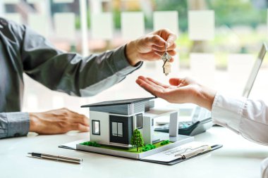 Emlakçı, alıcıya evin anahtarlarını modern minyatür ev modelinin olduğu bir masada veriyor..