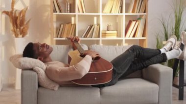 Gitar dinlendirici. Müzikal eğlence. Ev eğlencesi. Huzurlu hayalperest adam akustik telli enstrüman ile şarkı akorları çalıyor. Oturma odasındaki rahat kanepede hafta sonunun tadını çıkarıyor..