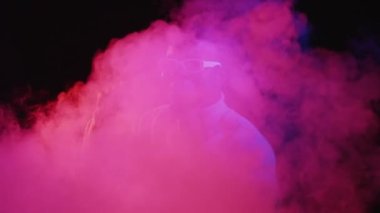 Gece kulübü dansı. Disko etkinliği. Parti eğlencesi. Tasasız, mutlu, stil sahibi genç adam canlı neon ışıklı duman efektiyle müzik ritminin tadını çıkarıyor..