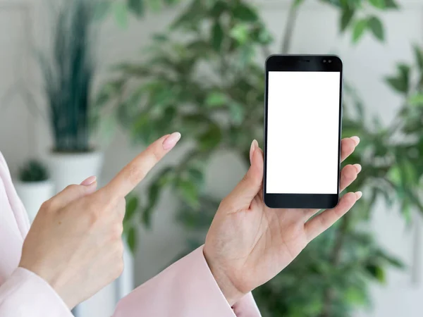 Mobile connection. Advertising mockup. Digital life. Female hands holding and pointing to smartphone with blank screen in light room interior.