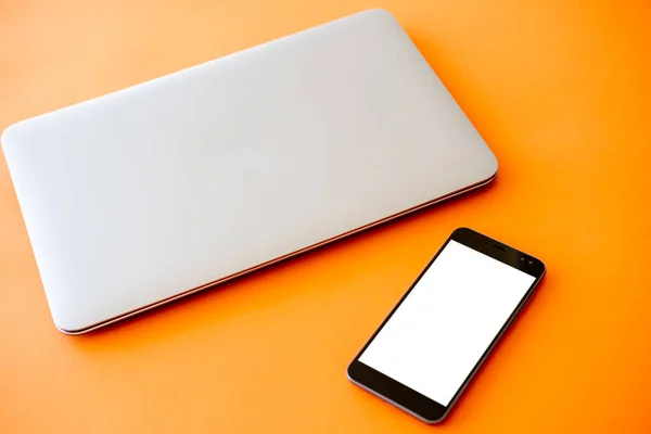 Digital mockup. Distance work. Online technology. Closed laptop and smartphone with white blank screen isolated orange.