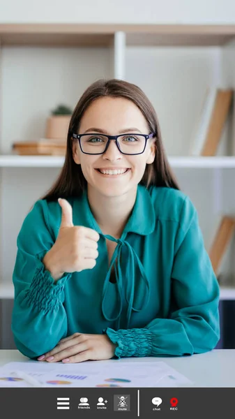 Online conference. Supporting woman. Screen mockup. Positive lady in spectacles sitting work desk showing like gesture in light room interior.