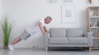 Koltuk egzersizi. Evde fitness. Sabah antrenmanı. Vücut heykeli. Güçlü orta yaşlı adam oturma odasının iç kısmında kanepe üzerinde şınav çekiyor..