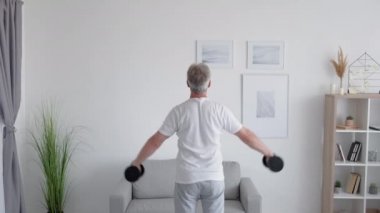 Kapalı spor salonu. Ev sporu. Aktif yaşam tarzı. Tanımlanamayan orta yaşlı bir adamın arka planda hafif oturma odasındaki dambıllarla kol kaslarını çalıştırması..