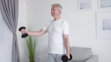 Evde fitness. Sabah egzersizi. Spor motivasyonu. Orta yaşlı ve esinlenilmiş bir adam oturma odasındaki halterli kol kaslarını çalıştırmaktan zevk alıyor..