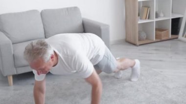 Sabah sporu. Evde eğitim. Vücut heykeli. Gülümseyen orta yaşlı, esinlenilmiş, orta yaşlı bir adam oturma odasının içinde yerde şınav çekiyor..