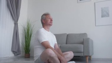 Evde yoga. Meditasyon terapisi. Pozitif yaşam tarzı. Mutlu, orta yaşlı, esinlenilmiş bir adam. Eğitimden zevk alıyor. Aydınlık bir iç mekanda bağdaş kurarak oturuyor..
