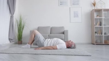 Evde fitness antrenmanı. Yoga pratiği. Sabah sporu. Rahat orta yaşlı bir adam oturma odasının iç kısmında yerde yatarak kalça kaldırma egzersizi yapıyor..