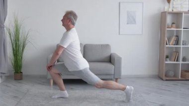 Kapalı alanda eğitim. Ev egzersizi. Fitness ısınıyor. Sabah sporu. Orta yaşlı bir adam aydınlık oturma odasında bacaklarını esnetiyor..