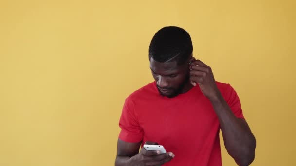 音楽アプリ Mp3プレイリスト 人を捕まえろ 現代の技術 オレンジのコピースペースに隔離された携帯電話で音を楽しむ無線イヤホンを身に着けている満足したアフリカの男背景 — ストック動画