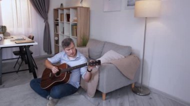 Müzikal hobi. Erkek oyuncu. Ev tatili. Sıradan orta yaşlı bir adam, yerde oturmuş, ışık odası iç açısında akustik gitar çalmaktan zevk alıyor..