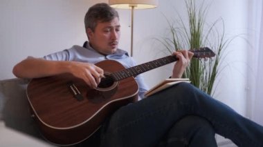 Müzik bestelemek. İlham verici bir adam. Ev tatili. Gündelik orta yaşlı bir adam kanepede oturup gitar çalıyor ve ışık odasının içindeki akorları not alıyor..