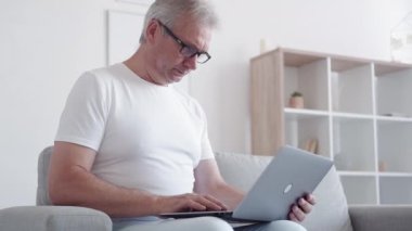 Dijital sorun. İnternet stresi. Sanal bir yanlış anlama. Hayal kırıklığına uğramış orta yaşlı bir adam evde çalışıyor. İnternette projeyi tartışıyor. Açık iç mekanda koltukta dizüstü bilgisayar kullanıyor..