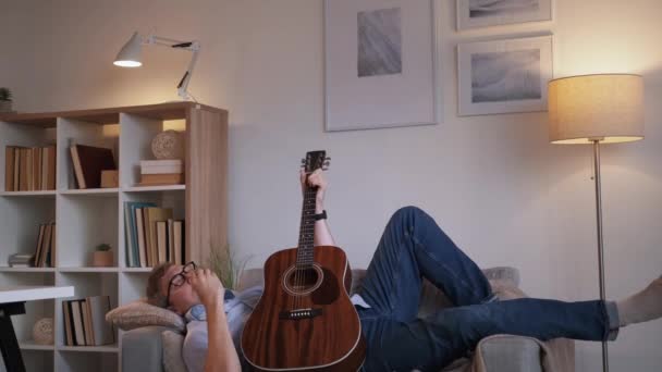 音乐灵魂 随便的男人 回家休息 受启发的休闲中年男人躺在沙发上弹奏吉他手的歌 在光线室的内部谱曲 — 图库视频影像