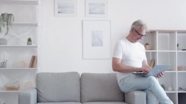 Çevrimiçi iletişim. Sanal sohbet. Sosyal medya. Rahat gülümseyen orta yaşlı bir adam açık modern ev iç mekanında dizüstü bilgisayarla mesaj yazıyor..
