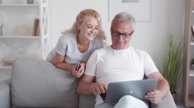 Aile aleti. Çevrimiçi ev eğlencesi. Bilgi teknolojisi. Gülümseyen orta yaşlı bir baba kızı hafif oturma odasındaki kanepede dizüstü bilgisayar kullanıyor..