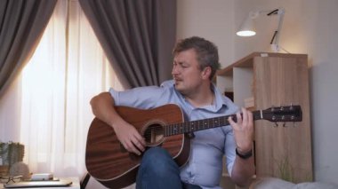 Müzik ilhamı. Mutlu adam. Ev tatili. Yakışıklı, orta yaşlı, rahat, rahat, koltukta oturup gitar çalan, aydınlık odada eğlenen bir adam..