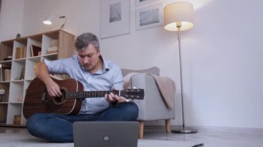 İnternetten ders. Erkek öğrenci. Uzaktan eğitim. Yakışıklı, orta yaşlı bir adam gitarla sanal konferans yapıyor. Dizüstü bilgisayarla zemin aydınlatma odasının iç tarafında oturuyor..