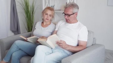 Aile hafta sonu eğlencesi. Ev tatili. Sanal eğlence. Sıkılmış orta yaşlı baba kitap okumayı bırakıp modern iç mekanlarda kanepede joysticklerle oyun oynuyor..