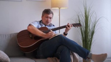 Müzik ilhamı. Erkek besteci. Ev tatili. Gündelik orta yaşlı bir adam koltukta oturup gitar çalıyor ve aydınlık odada yeni bir melodi için notalar alıyor..
